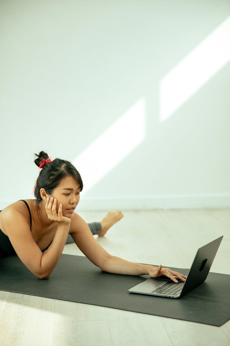 Side view of concentrated ethnic female in activewear stretching legs on mat on floor while browsing netbook in light room and looking at screen resting head on hand